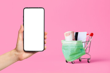 Virus protection and online purchase of medicines. A shopping cart with a medical mask on, filled with cash and a Bank card. A female's hand holds a smartphone. Pink background. Mock up