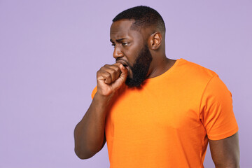 Sick young african american man wearing basic casual empty blank orange t-shirt standing coughing or sneezing covering mouth with hand isolated on pastel violet colour background studio portrait.