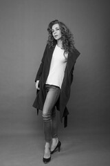 A beautiful slender brunette in a black coat, jeans and a white T-shirt stands in the studio on a gray background, bw