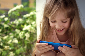 a little blonde girl enthusiastically plays games on the phone sitting on the windowsill against the background of a flowering tree