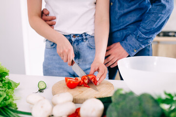 a man hugs a girl while she cuts a tomato on a salad.