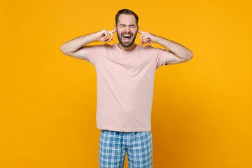 Crazy young man in pajamas home wear covering ears with fingers keeping eyes closed screaming while resting at home isolated on yellow background studio portrait. Relax good mood lifestyle concept.