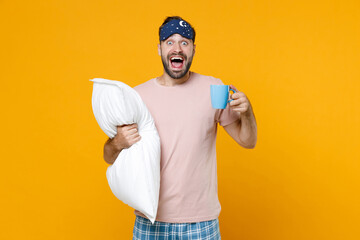 Surprised young bearded man 20s in pajamas home wear sleep mask hold pillow cup of coffee or tea...