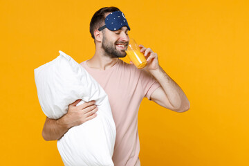 Naklejka premium Smiling young bearded man 20s in pajamas home wear sleep mask hold pillow glass of orange juice while resting at home isolated on yellow background studio portrait. Relax good mood lifestyle concept.