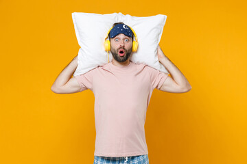 Shocked young bearded man 20s in pajamas home wear sleep mask hold pillow listening music with headphones while resting at home isolated on yellow background studio portrait. Relax good mood concept.
