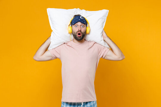 Shocked young bearded man 20s in pajamas home wear sleep mask hold pillow listening music with headphones while resting at home isolated on yellow background studio portrait. Relax good mood concept.