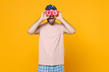 Surprised young man in pajamas home wear sleep mask imitating glasses or binoculars with donuts while resting at home isolated on yellow background studio portrait. Relax good mood lifestyle concept.