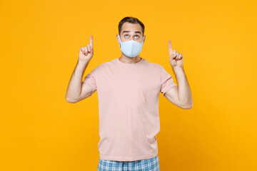 Young man in pajamas home wear face mask safe from coronavirus virus covid-19 pointing index fingers up on mock up copy space resting at home isolated on yellow background. Relax good mood concept.