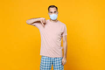 Dissatisfied displeased young man in pajamas home wear face mask safe from coronavirus virus covid-19 showing thumb down resting at home isolated on bright yellow background. Relax good mood concept.