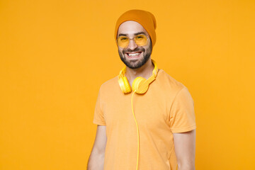 Obraz premium Smiling cheerful handsome attractive young bearded man 20s wearing basic casual t-shirt headphones eyeglasses hat standing looking camera isolated on bright yellow colour background, studio portrait.