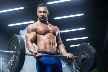 Fototapeta na wymiar Bottom up view of a muscular athlete lifting heavy barbell showing his muscles in a gym standing in smoke