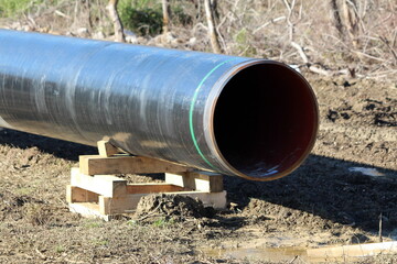 Large diameter pipe at local oil pipeline construction site supported with wooden supports on top of wet soil surrounded with dry vegetation and grass on cold sunny winter day