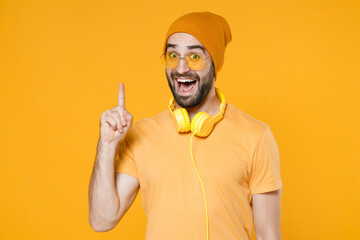Excited young bearded man in casual t-shirt headphones eyeglasses hat standing holding index finger...