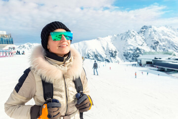 Fototapeta na wymiar Portrait of beautiful young adult caucasian woman in sport suit, hat, sunglasses and backpack looking and smiling on mountain peak covered with snow on bright sunny winter day at alpine ski resort