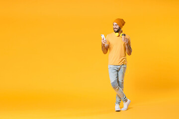 Full length of smiling young man wearing basic casual t-shirt headphones hat using mobile cell phone typing sms message hold paper cup of coffee or tea isolated on yellow background, studio portrait.