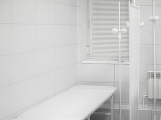 White medical couch for examining and treating a patient in light clean doctor's cabinet. Window with shutters, dripper stands, tiled walls. Doctor's office. Workplace