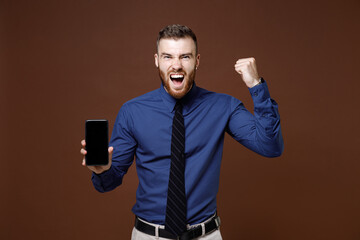 Happy young business man in blue shirt tie hold mobile cell phone with blank empty screen doing winner gesture isolated on brown background studio portrait. Achievement career wealth business concept.