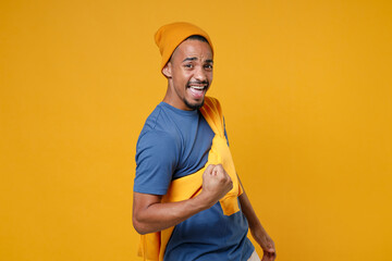 Joyful happy young african american man 20s in basic casual blue t-shirt hat standing clenching fists doing winner gesture looking camera isolated on bright yellow colour background, studio portrait.