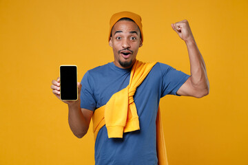 Amazed young african american man in blue t-shirt hat standing hold mobile cell phone with blank empty screen mock up copy space doing winner gesture isolated on yellow background, studio portrait.