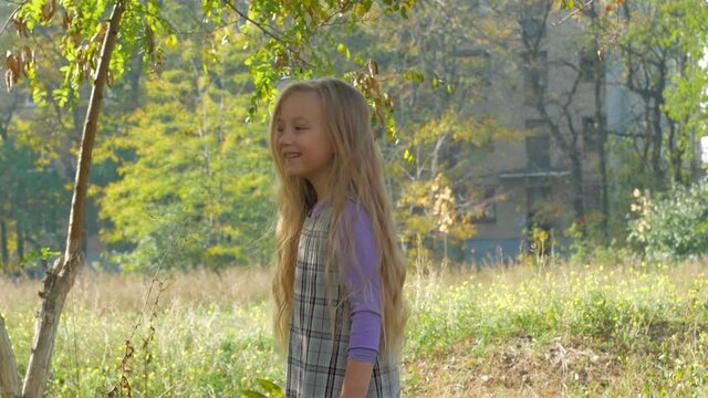 Beautiful girl with long blonde hair jumping on the nature. Smiling child plays in autumn park on a sunny day. Cute happy kid in dress walking alone dreaming.Green grass with yellow leaves outdoor