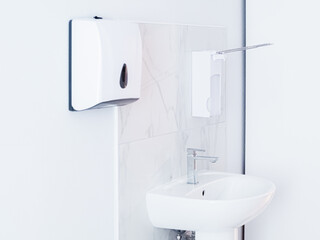 Bathroom with soap dispenser, white sink with metal faucet, and plastic tissue paper box, towel hanging on white wall. Light interior