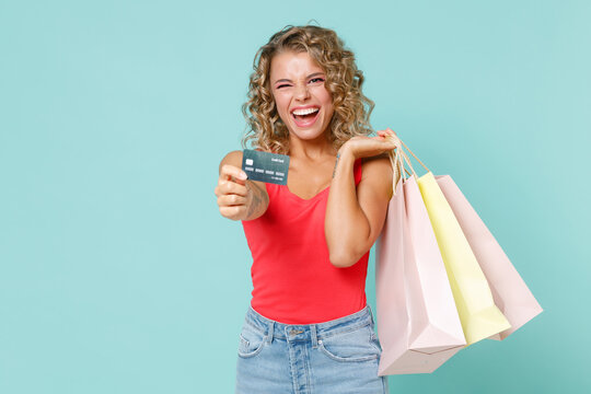 Funny blinking young blonde woman 20s wearing pink basic casual tank top hold package bags with purchases after shopping credit bank card isolated on blue turquoise colour background studio portrait.