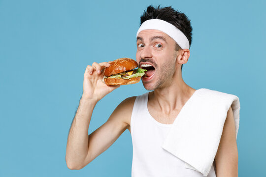 Amazed Young Sporty Fitness Man With Skinny Body Sportsman In Headband Shirt Towel Hold Eating American Classic Fast Food Burger Isolated On Blue Background. Workout Gym Sport Motivation Concept.