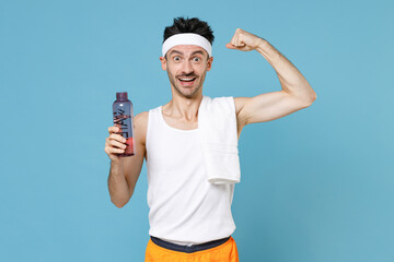 Excited young fitness man with thin skinny body sportsman in headband shirt shorts hold bottle of water showing biceps muscles isolated on blue background studio. Workout gym sport motivation concept.