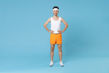 Fototapeta na wymiar Full length portrait smiling young fitness man with skinny body sportsman in headband shirt shorts standing with arms akimbo on waist isolated on blue background. Workout gym sport motivation concept.