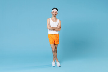 Fototapeta na wymiar Full length portrait smiling young sporty fitness man with skinny body sportsman in white headband shirt shorts holding hands crossed isolated on blue background. Workout gym sport motivation concept.