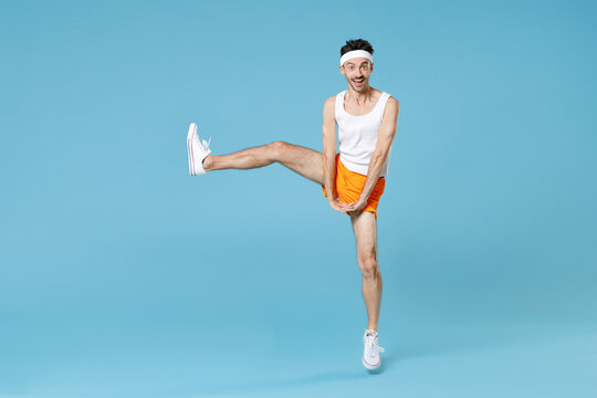 Full Length Portrait Of Cheerful Young Sporty Fitness Man With Skinny Body Sportsman In White Headband Shirt Shorts Rising Leg Isolated On Blue Background Studio. Workout Gym Sport Motivation Concept.