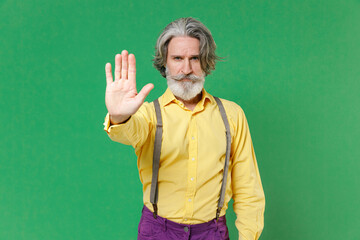 Displeased elderly gray-haired mustache bearded man wearing casual yellow shirt suspenders showing STOP gesture with palm looking camera isolated on bright green colour background, studio portrait.