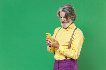 Smiling handsome elderly gray-haired mustache bearded man wearing casual yellow shirt suspenders using mobile cell phone typing sms message isolated on bright green colour background studio portrait.