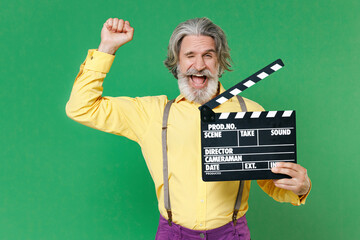 Blinking elderly gray-haired mustache bearded man wearing yellow shirt suspenders hold classic black film making clapperboard doing winner gesture isolated on green colour background, studio portrait.