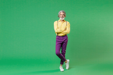 Full length of smiling elderly gray-haired mustache bearded man wearing casual yellow shirt suspenders holding hands crossed looking camera isolated on bright green colour background, studio portrait.