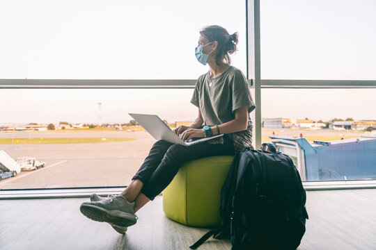 A young woman in a protective mask on her face sits with a laptop in the departure area of the airport and is waiting for her flight. Travel to pandemic covid coronavirus, new normal lifestyle