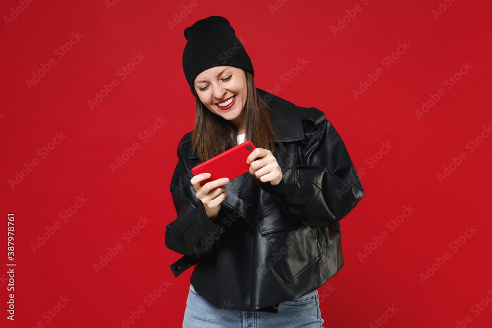 Wall mural Smiling cheerful funny young brunette woman 20s wearing basic casual black leather jacket white t-shirt hat playing game on mobile cell phone isolated on bright red colour background studio portrait.