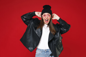 Excited surprised young brunette woman 20s in casual black leather jacket white t-shirt hat eyeglasses put hands on head keeping mouth open isolated on bright red colour background studio portrait.