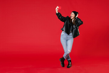 Full length of excited cheerful young brunette woman 20s wearing black leather jacket white t-shirt hat doing selfie shot on mobile phone put hand on head isolated on red background studio portrait.