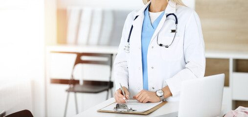 Unknown woman-doctor at work in sunny clinic. Female physician controls medication history record, close-up. Medicine concept