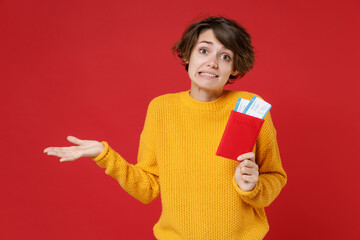 Confused young traveler tourist woman in yellow sweater hold passport tickets spreading hands isolated on red background. Passenger traveling abroad on weekends getaway. Air flight journey concept.