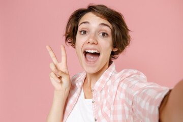 Close up of excited young brunette woman wearing casual checkered shirt standing doing selfie shot on mobile phone showing victory sign isolated on pastel pink colour background, studio portrait.