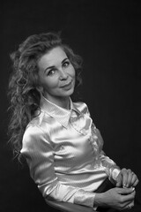 A beautiful slender brunette with long curly hair in a white satin blouse sits in the studio on a black background, bw