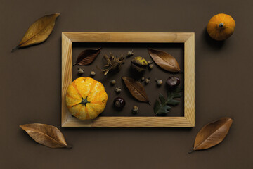 Autumn composition made of pumpkins, dried leaves, chestnuts and acorns in a wooden frame on brown background.