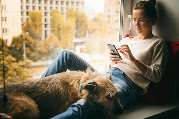 A young woman with her brown dog is sitting on a windowsill overlooking the city, looking at the...