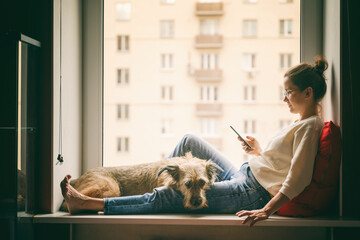 A young woman with her brown dog is sitting on a windowsill overlooking the city, looking at the smartphone screen and stroking her pet