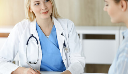 Woman-doctor and patient sitting and talking in sunny clinic. Blonde therapist is cheerfully smiling