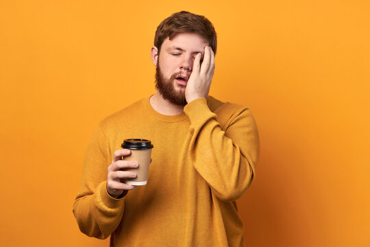 Close up portrait of man on isolated yellow studio background, taking coffee in takeaway paper cup, posing with closed eyes and open mouth, holding hand on cheek