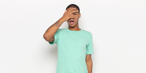 young black man looking shocked, scared or terrified, covering face with hand and peeking between...