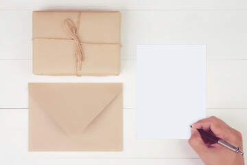 Hand of man writing letter with blank greeting card and having gift box with kraft paper on wooden table, present or package, postcard mockup, top view, flat lay, copy space, holiday concept.